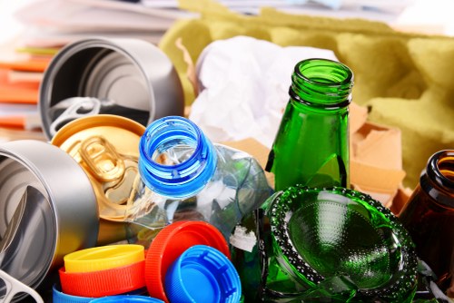 A close-up view of various used glass bottles and plastic containers scattered on a surface, including a green glass bottle standing upright, a blue plastic bottle with its cap removed, and a transparent plastic bottle with a golden cap, alongside crushed aluminium cans with orange and yellow caps. In the background, there are soft, crumpled packing materials and a yellow fabric or foam padding, indicating a collection of recyclable waste. The scene suggests an outdoor or possibly indoor area dedicated to waste collection, typical of rubbish removal services operating in central London areas such as Soho. The image emphasizes the sorting and disposal of different packaging materials, reflecting the work of a professional waste management company like Commercial Waste Soho, which offers rubbish removal services in the local postcode district, contributing to responsible waste recycling and clearance efforts in the region.