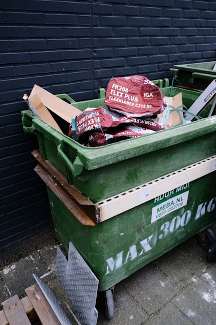 A green plastic rubbish bin positioned outdoors against a dark brick wall, filled with red and black flexible garden hose packaging and related materials. The bin is situated on a paved surface and re