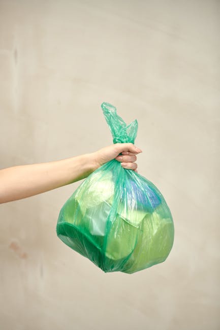 A green plastic rubbish bin positioned outdoors against a dark brick wall, filled with red and black flexible garden hose packaging and related materials. The bin is situated on a paved surface and re